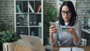 Ofis masasında kredi kartı ve akıllı telefonunu kontrol eden kişi; izinsiz kart işlemleri ve harcama itirazı rehberi için temsili fotoğraf