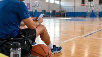Spor salonunda basketbol topu ve antrenman çantası yanında telefonuyla işlem yapan sporcu